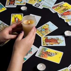 May include: A lit candle in a glass holder is held over a spread of tarot cards on a black surface. Several tarot cards are arranged around the candle, with additional candles in the background. The cards feature colorful illustrations and text.