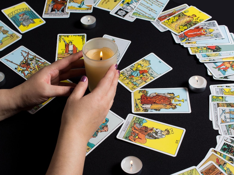 May include: A lit candle in a glass holder is held over a spread of tarot cards on a black surface. Several tarot cards are arranged around the candle, with additional candles in the background. The cards feature colorful illustrations and text.