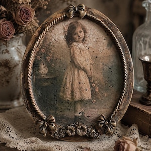 Peut inclure: Un cadre photo ovale avec une finition dorée vieillie, doté d'une bordure en corde torsadée et de nœuds décoratifs en haut et en bas. Le cadre contient une image sépia d'un enfant en robe, avec une texture craquelée visible.