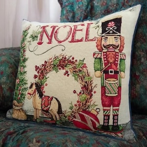 Puede incluir: Cojín decorativo navideño con un diseño festivo. Presenta la palabra "NOEL" en rojo, un soldado cascanueces, una corona, un caballito balancín y un adorno rojo. El cojín tiene un borde azul.