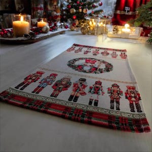 May include: A rectangular Christmas table runner featuring a repeating pattern of toy soldier nutcrackers in various colors. The runner has a cream-colored background with a red and green plaid border and a central wreath design.