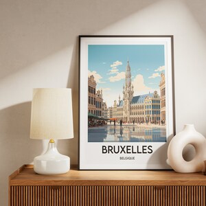 May include: A framed print of a colorful illustration of the Grand Place in Brussels, Belgium. The print features a view of the city square with the Town Hall in the center. The text "BRUXELLES BELGIQUE" is printed below the image.