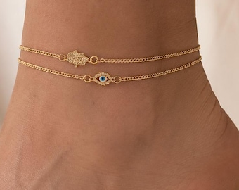 Könnte beinhalten: Eine goldfarbene Fußkette mit zwei Strängen und zartem Kettendesign. Ein Strang zeigt einen kleinen, schimmernden Hamsa-Hand-Anhänger, während der andere Strang einen runden Nazar-Auge-Anhänger mit einem zentralen blau-weißen Glasdetail aufweist.