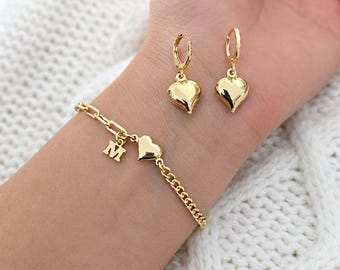 May include: A gold-coloured jewellery set comprising a bracelet with a heart charm and an initial "M" charm, plus matching heart-shaped hoop earrings. The bracelet has a chain link design. The earrings and bracelet are displayed on a wrist.