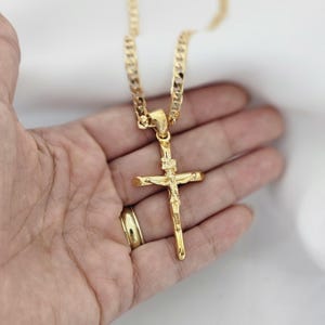 Puede incluir: Collar con colgante de cruz de color dorado. El colgante presenta una representación detallada de Jesús en la cruz. El collar tiene un diseño de cadena de bordillo. La joyería se muestra sobre un fondo neutro.