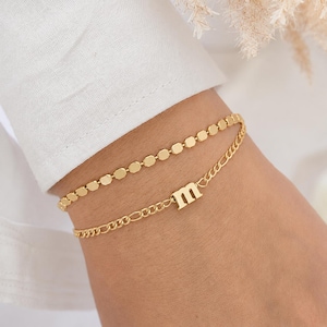 Könnte beinhalten: Ein goldfarbenes, zweireihiges Armband bestehend aus einer Kette mit kleinen flachen rechteckigen Gliedern und einer zweiten Kette mit klassischen Gliedern, akzentuiert durch einen kleinen Buchstaben m als Anhänger.