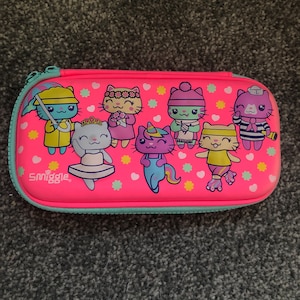 May include: Pink pencil case with a zip closure and a Smiggle logo. The case features a colourful design of cartoon cats with different accessories and expressions.