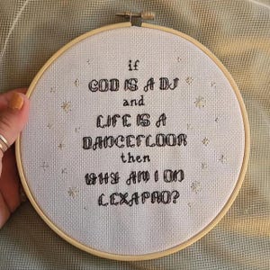 Könnte beinhalten: Ein Kreuzstich-Stickrahmen mit dem Text "If God is a DJ and Life is a Dancefloor then Why am I on Lexapro?" in schwarzem Faden. Die Stickerei befindet sich auf weißem Stoff, umgeben von einem Holzrahmen.
