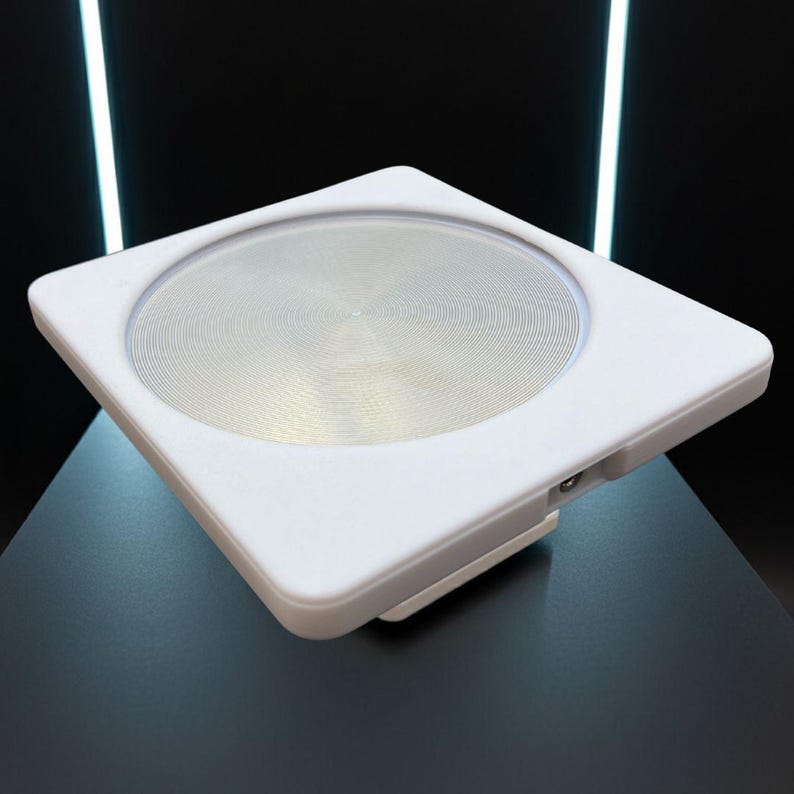 May include: A white, square device with a circular, metallic centre. The device has a raised edge and a small port on the side. The background is black with two vertical white lights.