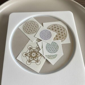 May include: A selection of small, square stickers with geometric patterns, including the Flower of Life and Metatron's Cube. The stickers, in gold, purple, and green, are arranged on a white square tray with a circular, textured base.