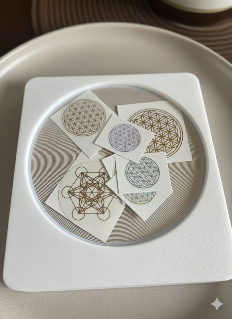 May include: A selection of small, square stickers with geometric patterns, including the Flower of Life and Metatron's Cube. The stickers, in gold, purple, and green, are arranged on a white square tray with a circular, textured base.