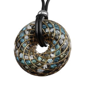 May include: A pendant necklace featuring a circular, open-center design. The pendant is a mosaic of blue, black, and clear stones set within a gold-colored metal frame. The necklace has a black cord with a silver-colored clasp.