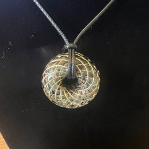 Peut inclure: Un collier en or et noir avec un pendentif géométrique. Le pendentif est fait de fil et a un design en spirale. Le collier est sur un fond noir.