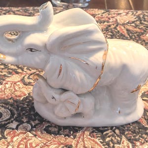 May include: White ceramic elephant figurine with gold accents. The elephant is depicted with a baby elephant at its feet. The figurine has detailed features and is a decorative item.