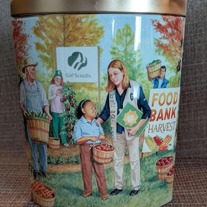 May include: A decorative tin with a gold lid and a painted scene of Girl Scouts harvesting food for a food bank. The artwork features people carrying baskets of produce and the words "Food Bank Harvest."