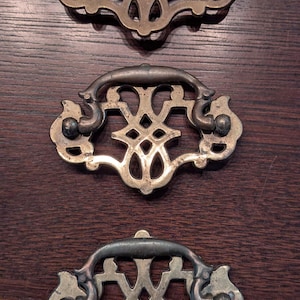 May include: Three antique brass drawer pulls with ornate, symmetrical designs. Each pull features a curved handle and decorative backplates with intricate cutouts and screw attachments. The pulls are set against a dark wood surface.