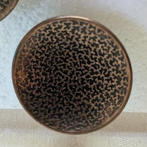 May include: Close-up of a copper-colored, round knob with a textured, speckled surface. The knob has a dark brown pattern and a slightly raised edge. The knob is likely for a cabinet or drawer.