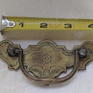 May include: Antique brass drawer pull with a floral design. The pull measures approximately 4 inches in length. The handle is curved and attached to a decorative backplate with engraved details. The pull has a vintage, aged appearance.