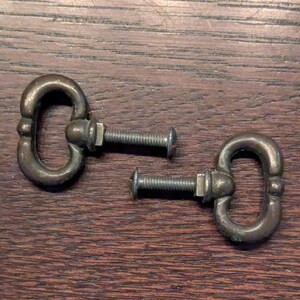 May include: Two antique brass drawer pulls with oval handles and threaded bolts. The hardware is aged with a patina, suggesting vintage or repurposed use. The pulls are set against a dark wood surface.
