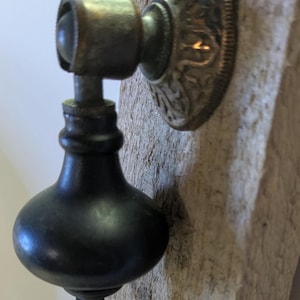 May include: Close-up of an antique brass and black door pull. The pull features a decorative, circular backplate with an ornate design. The handle is a dark, bulbous shape, and the hardware is mounted on a weathered wooden surface.