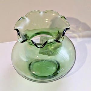 May include: A green glass vase with a ruffled rim and a round body. The rim has a black trim. The vase is made of transparent glass, allowing light to pass through. The vase is likely used for decorative purposes.