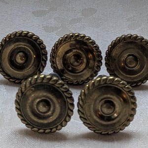 May include: Five antique brass drawer pulls with a rope-like edge design. Each pull has a circular face with a central indentation and a raised outer ring. The pulls are arranged on a white, textured surface, showcasing their vintage aesthetic.
