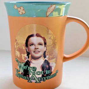 May include: Orange ceramic mug featuring a portrait of Judy Garland as Dorothy from The Wizard of Oz. The mug has a teal rim with decorative elements and the quote "We're Not in Kansas Anymore."