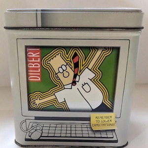 May include: A rectangular tin featuring a Dilbert cartoon character inside a computer screen. The character is wearing glasses and a tie. The tin has the word "DILBERT" on the side and a sticky note that says "REMEMBER TO LOWER EXPECTATIONS!"