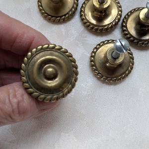 Puede incluir: Pomos de armario antiguos de lat&oacute;n con un dise&ntilde;o de borde similar a una cuerda. Los pomos tienen una hendidura circular central y est&aacute;n unidos a tornillos. Varios pomos son visibles, junto con tornillos sueltos.