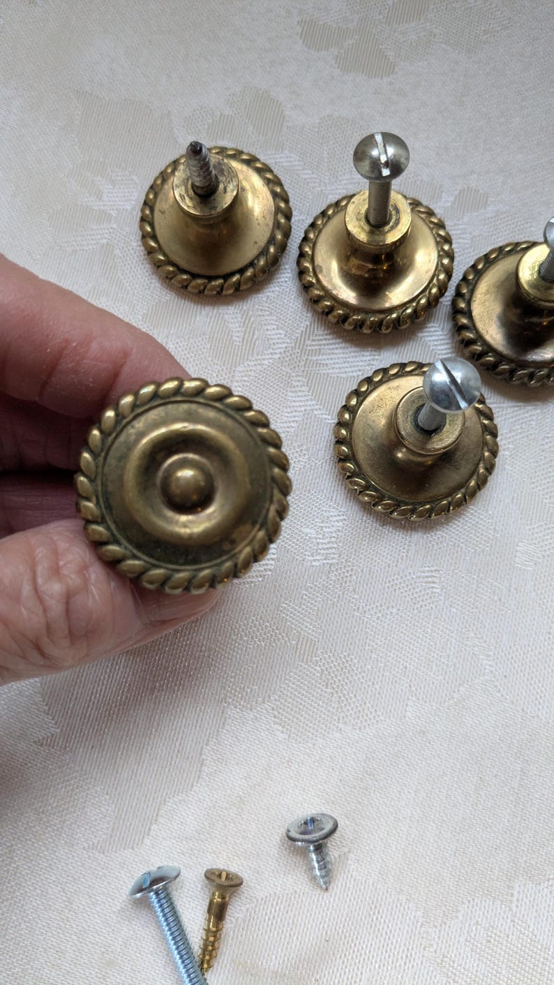 Puede incluir: Pomos de armario antiguos de lat&oacute;n con un dise&ntilde;o de borde similar a una cuerda. Los pomos tienen una hendidura circular central y est&aacute;n unidos a tornillos. Varios pomos son visibles, junto con tornillos sueltos.