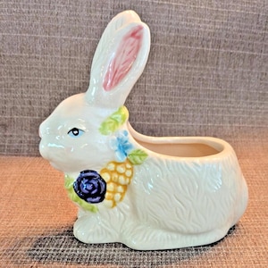 May include: A ceramic bunny planter in cream with a textured surface. The bunny has pink inner ears, blue eyes, and is adorned with a floral arrangement of blue, yellow, and purple flowers. The planter is designed to hold small plants or flowers.