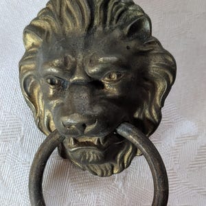May include: Antique brass lion head door knocker with a ring handle. The lion's face has detailed features, including a mane, eyes, nose, and mouth. The knocker is made of metal and has a vintage appearance.