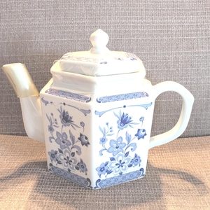 May include: White porcelain teapot with a square body and a lid. The teapot is decorated with blue floral patterns and trim. It has a curved handle and a spout with a gold-colored tip. A decorative piece for the kitchen.