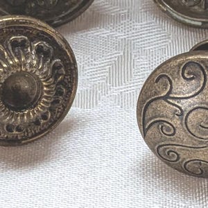 May include: Close-up of several antique brass cabinet knobs. The knobs have intricate designs, including floral and scroll patterns. The knobs are round and have a tarnished, vintage appearance.