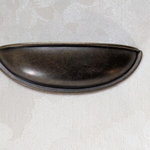 May include: A bronze-toned, half-moon shaped drawer pull. The hardware has a curved design with a slightly raised lip around the edges. The finish has a vintage, antique appearance, suggesting a classic or retro style.