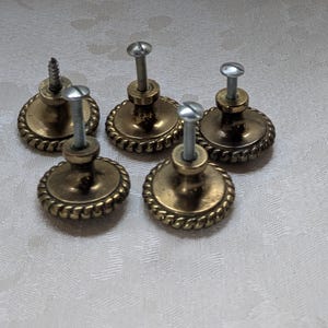 Puede incluir: Cinco tiradores de caj&oacute;n antiguos de lat&oacute;n con un dise&ntilde;o de borde similar a una cuerda. Cada tirador tiene una cara redonda, ligeramente abovedada y un tornillo para la fijaci&oacute;n. El lat&oacute;n tiene una p&aacute;tina c&aacute;lida y envejecida, que sugiere un estilo vintage o retro.