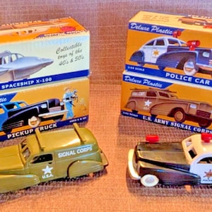 May include: Vintage toy cars and boxes. The image shows four toy cars, including a pickup truck, police car, and a U.S. Army Signal Corps vehicle. The boxes feature illustrations of the toys and text that reads "Deluxe Plastic".