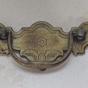 May include: Antique brass drawer pull with a floral design. The pull features a curved handle and decorative end pieces. The center of the pull has a flower and leaf design. The hardware is mounted with two small screws.