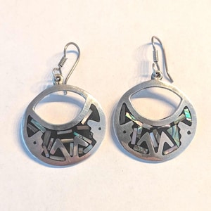 May include: Pair of silver-tone earrings with a circular design. Each earring features a cutout shape and is inlaid with iridescent abalone shell pieces. The earrings have a hook closure for pierced ears.