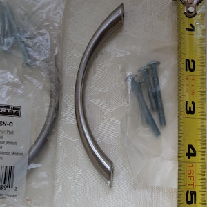 May include: Brushed nickel cabinet pulls with mounting screws. The curved pulls are approximately 4 inches long. A yellow tape measure shows the length in inches. Hardware for home improvement projects.
