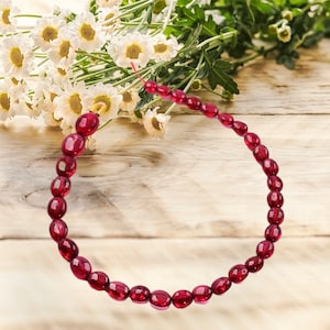 May include: A strand of deep red oval-shaped beads strung together to form a bracelet. The bracelet is lying on a wooden surface.