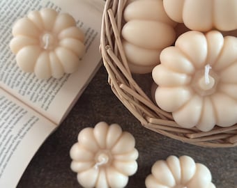 May include: A collection of ivory-colored pumpkin-shaped candles. Some candles are in a woven basket, while others are arranged on a wooden surface next to an open book. The candles have a realistic pumpkin design with a wick.
