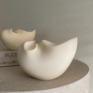 Könnte beinhalten: Zwei weiße, muschelförmige Kerzen, eine größer als die andere, mit einer strukturierten, gerippten Oberfläche. Die Kerzen stehen auf einer hellbraunen Oberfläche.