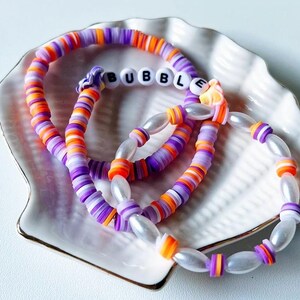 以下が含まれることがあります： 白い文字で「BUBBLE」と綴られたブレスレットを含む、カラフルなビーズのブレスレット3本。ブレスレットは白い貝殻の形をした皿に置かれています。
