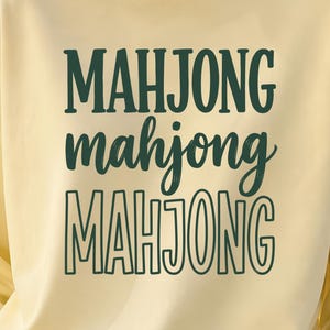 Op de afbeelding: Een lichtgeel sweatshirt met ronde hals waarop het woord MAHJONG in het donkergroen is gedrukt in drie verschillende lettertypes die verticaal zijn gestapeld. De bovenste regel is in een vet schreeflettertype, het midden in cursief en de onderste in omtrek.