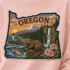 May include: A light pink crewneck sweatshirt featuring a large rectangular graphic of the state of Oregon. The design includes a beaver, a leaping fish, a lighthouse, a mountain, a waterfall, and pink flowers with the text OREGON at the top.