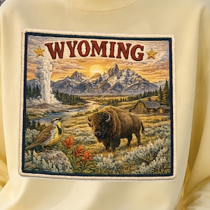 May include: A pale yellow sweatshirt featuring a rectangular graphic print of a Wyoming landscape. The design includes a bison, a bird, mountains, a geyser, and a cabin under a sunset sky with the text WYOMING flanked by two gold stars.