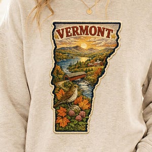May include: A beige crewneck sweatshirt featuring a central graphic shaped like the state of Vermont. The design depicts a landscape with mountains, a river, a covered bridge, a bird, and autumn leaves, with the word VERMONT printed at the top.