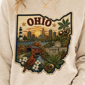 Puede incluir: Una sudadera beige de cuello redondo con un gráfico grande y detallado con la forma del estado de Ohio. El diseño incluye un horizonte urbano, un faro, un puente cubierto, un edificio del capitolio, un cardenal rojo y hojas de castaño de Indias.