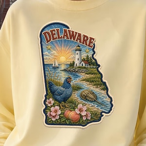 Könnte beinhalten: Ein blassgelbes Sweatshirt mit Rundhalsausschnitt und einem großen, staatsförmigen Grafikaufnäher von Delaware. Das Design zeigt einen Leuchtturm, ein Segelboot, ein blaues Huhn, einen Pfeilschwanzkrebs, Pfirsichblüten und einen Pfirsich.